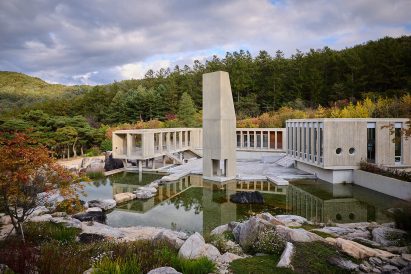 Large concrete complex with geometric forms reflected in a still pond, framed by forested hills