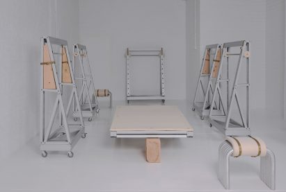 Minimalist industrial furniture installation featuring steel and wood modular frames on caster wheels in a white gallery space, arranged symmetrically around a central wooden platform.