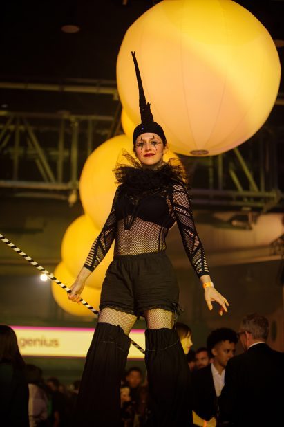 A stiltwalker wearing a black circus costume with a tall pointed headpiece, mesh top, ruffled collar, and voluminous leg coverings poses under large glowing yellow globe lights at a busy indoor event.