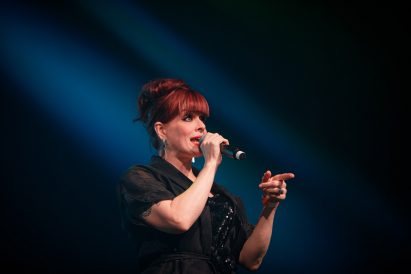 Ana Matronic standing on stage wearing a black sequin dress, illuminated by spotlights, holding a mic as she addresses the audience