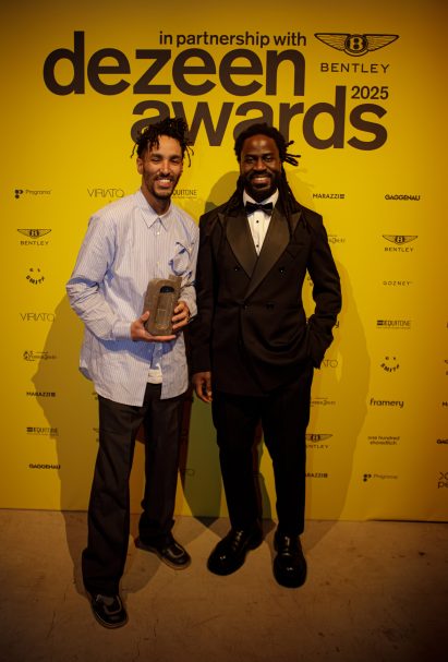 Andu Masebo and Tejumola Butler Adenuga posing in front of a bright yellow Dezeen Awards 2025 photo wall, one holding trophy, surrounded by sponsor logos