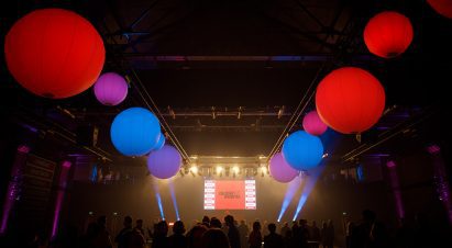 A crowd stands underneath bright colourful globe lights hanging from the ceiling, a screen with the Dezeen Awards logo in red is on the stage with strobes lights either side