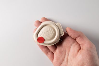 Photo of a hand holding a fish-shaped beige-coloured plant-pulp packaging design in the shape of a fish with a small red circular sticker stuck on it
