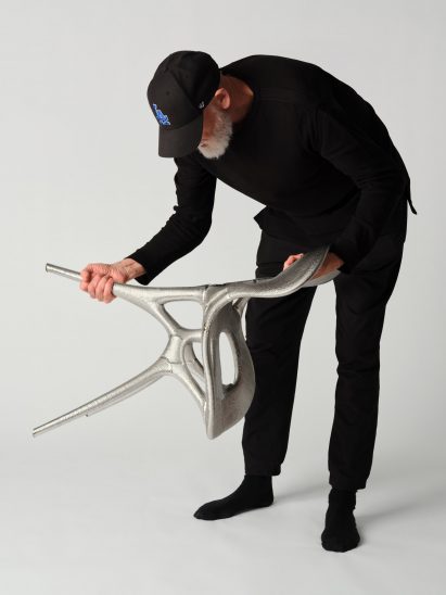 Photo of Ross Lovegrove handling the metal chair he co-created with Google DeepMind and Modem