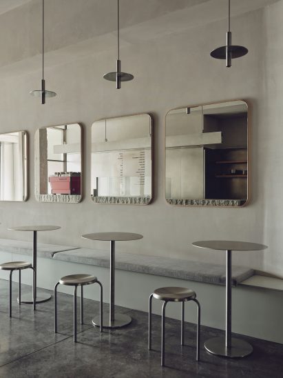 Minimalist cafe seating area with stainless steel tables and stools, bench seating, large rectangular wall mirrors, and custom pendant lights.