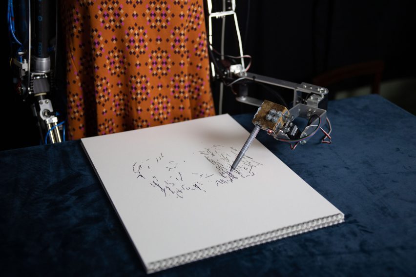 Photo of a drawing pad with a robot arm hovering above it holding a pen and drawing disjointed black lines