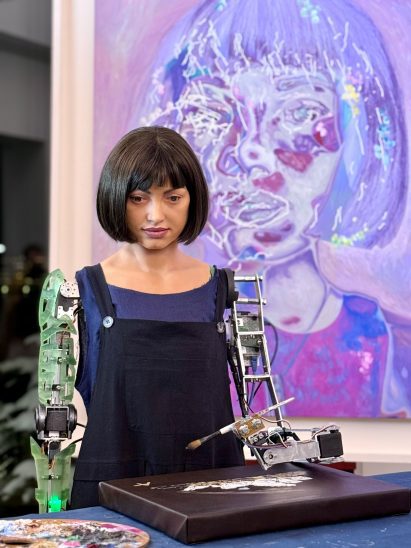 Photo of the Ai-Da humanoid robot with a paintbrush in her mechanised arm and an artwork in front of her and on the wall behind