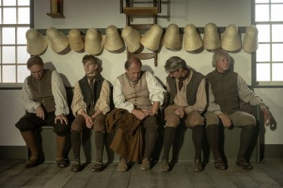 Photo of a scene in The Testament of Ann Lee with men sitting on bench seating below a peg board hanging hats and furniture