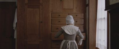 Still from The Testament of Ann Lee showing the back of a bonneted woman as she pulls out a drawer in wall-to-wall Shaker cabinetry