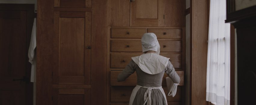 Still from The Testament of Ann Lee showing the back of a bonneted woman as she pulls out a drawer in wall-to-wall Shaker cabinetry