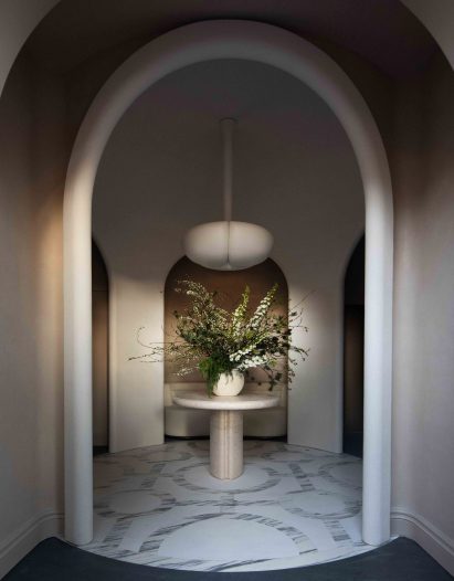 A domed room with a white archways arranged in a circular formation. In the middle is a circular table with a cylindrical base. On top of the table is a large arrangement of flowers. A large white lampshade is hanging from the ceiling.