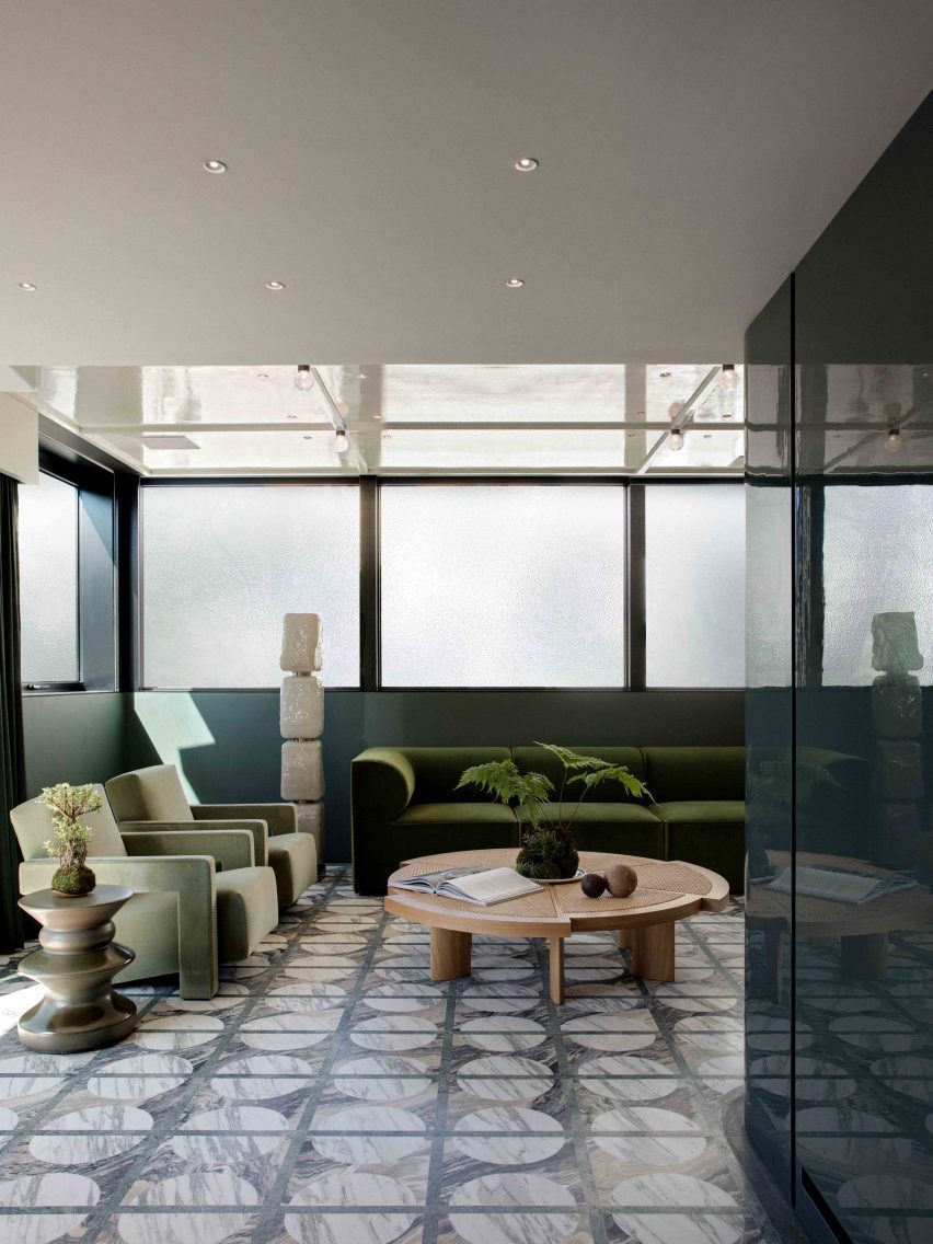 The top floor entertainment room shows patterned grey tiles, a wooden coffee table and a set of velvet green furnishings. Glass windows wrap around the room, and small stone sculptures stand between the sofas and armchairs.