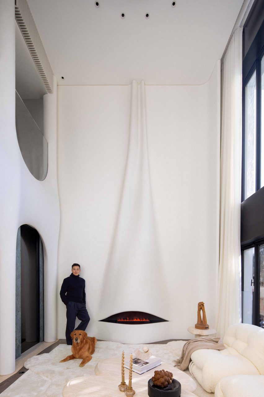 A tall, double-height fireplace that is integrated into a wall that has been carved to resemble a piece of draped cloth. The main architect, Nelson Chow is standing on the left of the fireplace with a dog laid on the floor beside him.