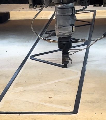 Photo of a large-scale 3D-printing nozzle extruding a dark plastic-glass composite material into a zigzag shape