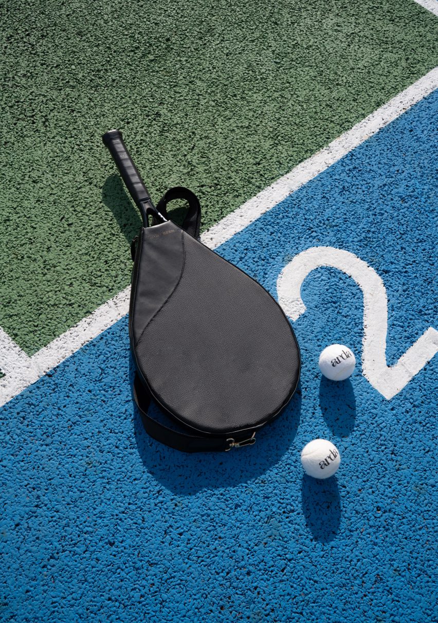 Photo of a tennis racquet in a black leather-like cover, lying on a tennis court beside two white tennis balls that say Arda