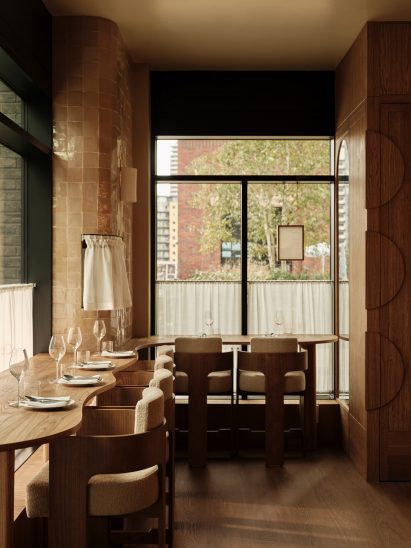 Nora restaurant interior with zellige tile clad walls, set dining tables with wine glasses, chairs, curved wooden tabletops and windows looking out to street level in an upscale dining space