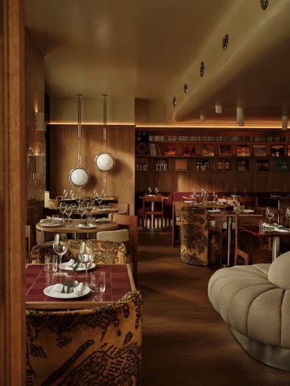 Nora restaurant interior featuring wood-panelled walls, sculptural pendant lights, vinyl record shelving, set dining tables with wine glasses, patterned upholstered chairs, tiled tabletops, red flooring, and cosy ambient lighting in an upscale dining space