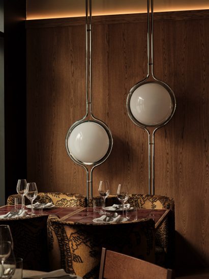 Nora restaurant interior featuring bespoke sculptural lighting , set dining tables with wine glasses, patterned upholstered chairs, tiled tabletops, red flooring, in an upscale dining space