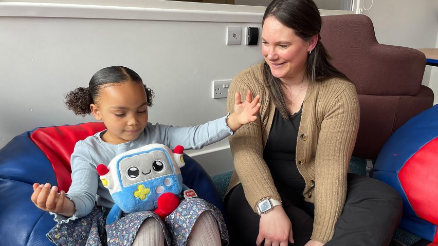 A child throwsher arms around the cuddly toy Gabbo while her mum watches from beside her