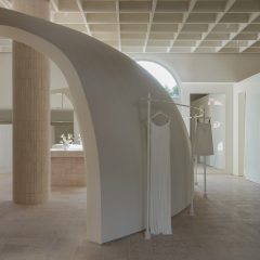 Image of a truncated domed partition wall with arched walkways connecting the main shop floor from a gloss-finished block of changing rooms at the far right of the image. A clothes rail with a hanging dress and skirt wraps around the dome's exterior.