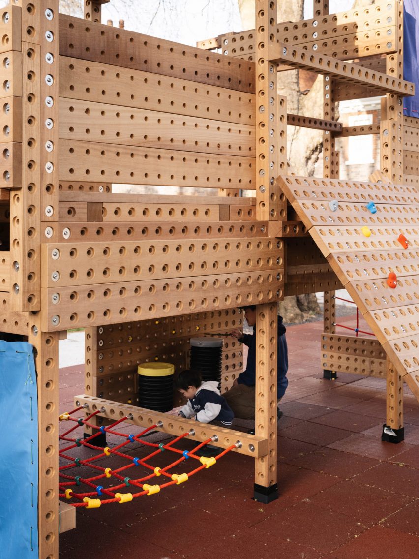 Photo of a two-storey wooden playground structure with children playing in the cubby-like space underneath the platform, with a bright red climbing net strung up between beams beside them