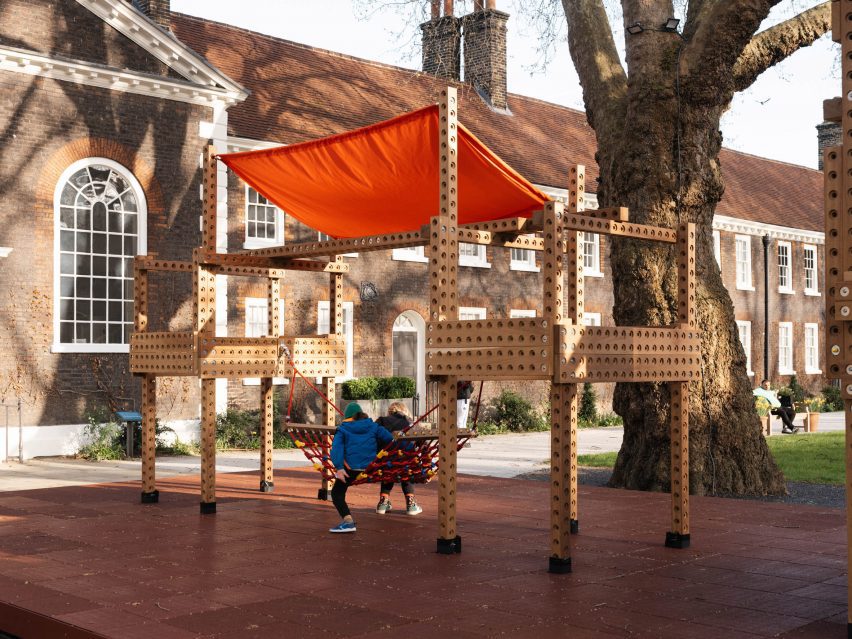 Photo of two kids sitting on a bright red hammock-like net strung up between beams on a wooden playground structure