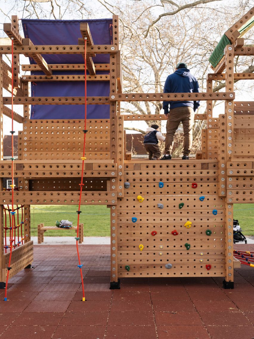 Photo of a two-storey wooden playground structure with a plank of colourful climbing holds going up to the top level and two red ropes with coloured footholds coming down like fireman's poles