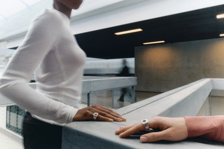 Photo of a woman's torso with her hand, wearing a chunky silver Quartz ring, resting on a low wall within an office-like building. Another hand with a Quartz ring is in the foreground