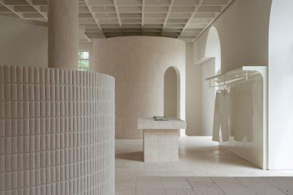 An image of a neutral, cream-toned interior with architectural elements including columns, a cylindrical room and a rectangular table made from travertine and limestone. On the right is a hanging rail with a pair of trousers on it.