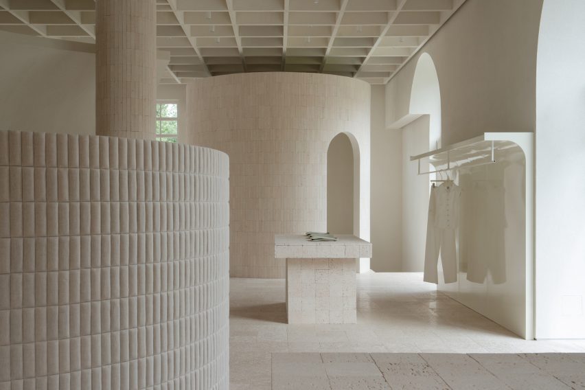 An image of a neutral, cream-toned interior with architectural elements including columns, a cylindrical room and a rectangular table made from travertine and limestone. On the right is a hanging rail with a pair of trousers on it.