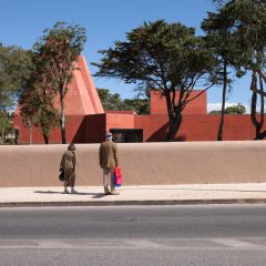 Street view of the museum behind a low wall with visitors looking towards the red concrete building
