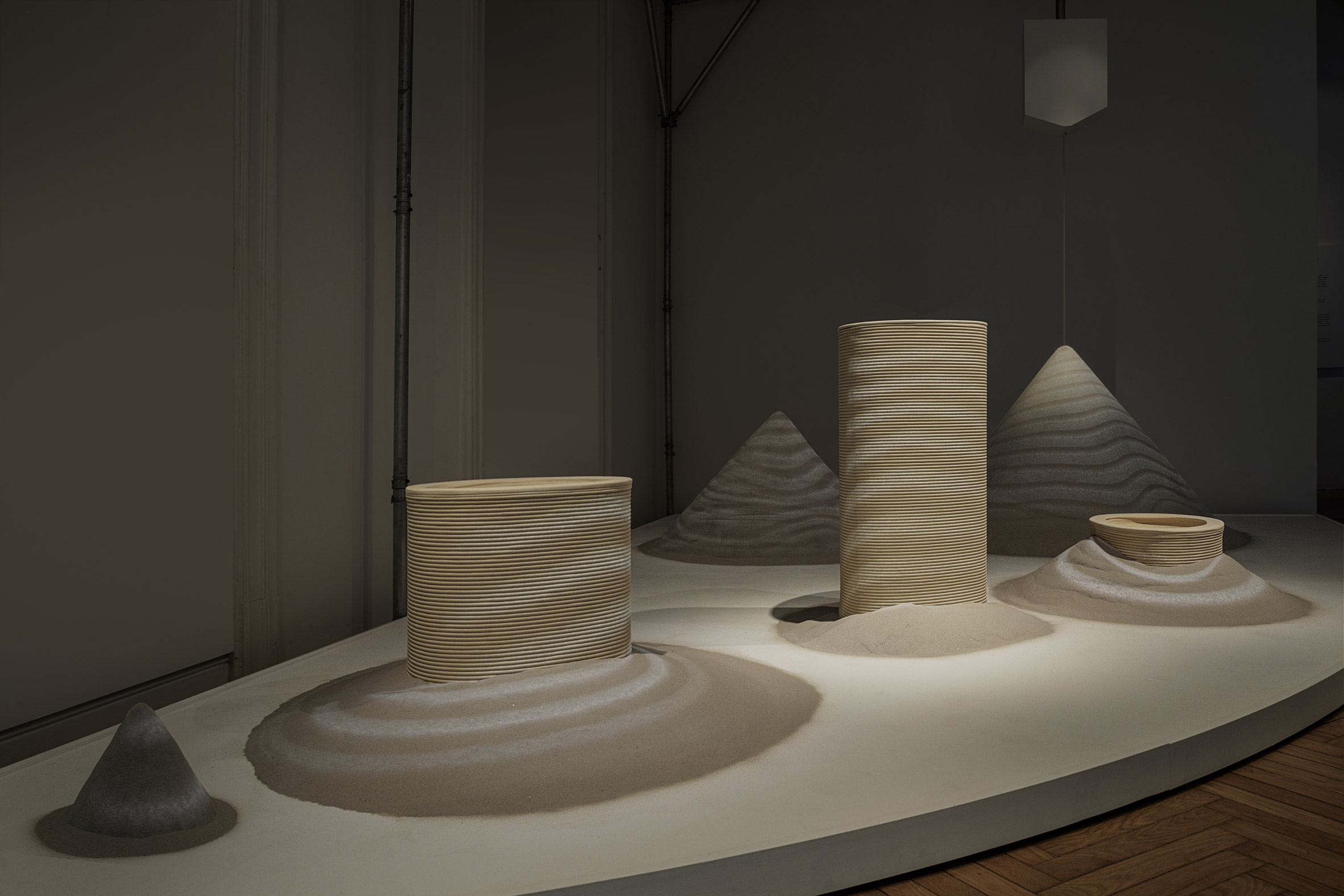 Layered cylindrical and conical forms made of ceramic sit in sand mounds beneath soft lighting