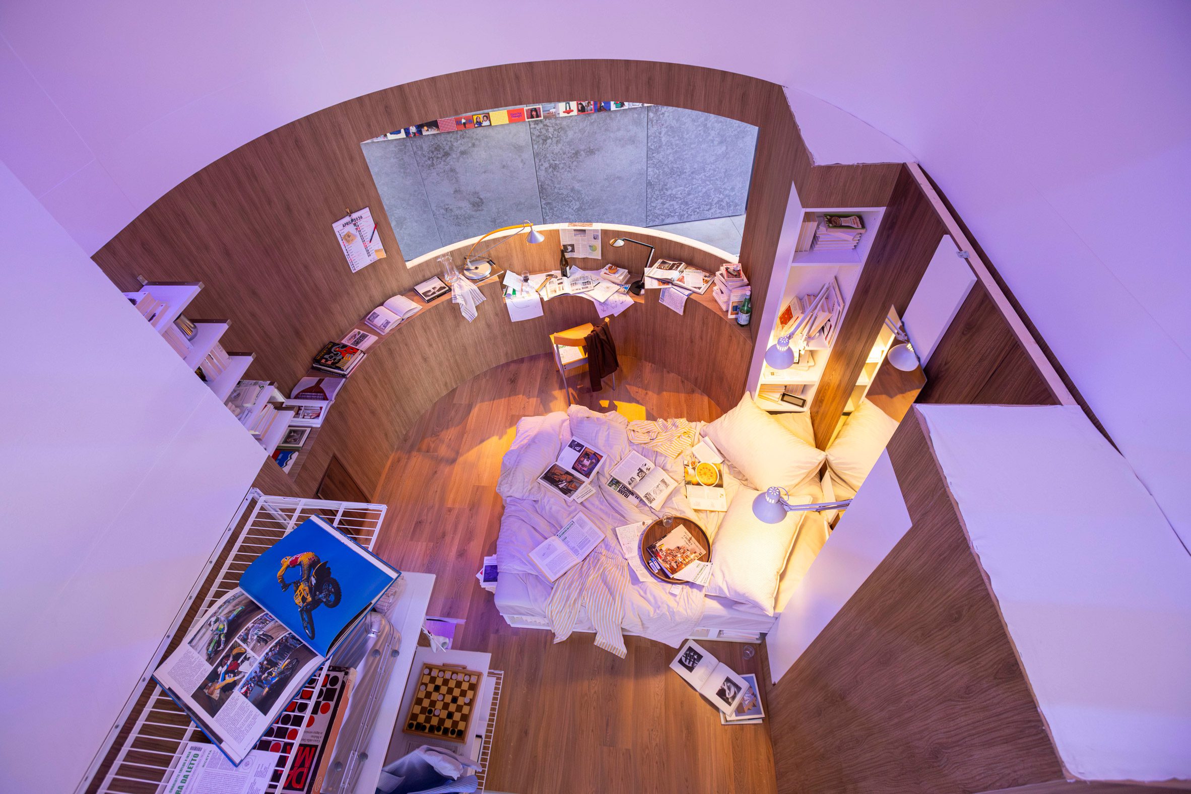 A compact bedroom with a curved timber desk, built-in shelving and a bed in the IKEA Food for Thought exhibition in Milan