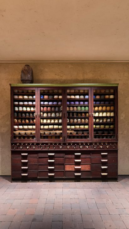 Photo of an antique apothecary cabinet in dark wood holding dozens of similar cups and bowls, with a gentle rainbow of glazes in the Chawan Cabinet exhibition at Prada Home Milan