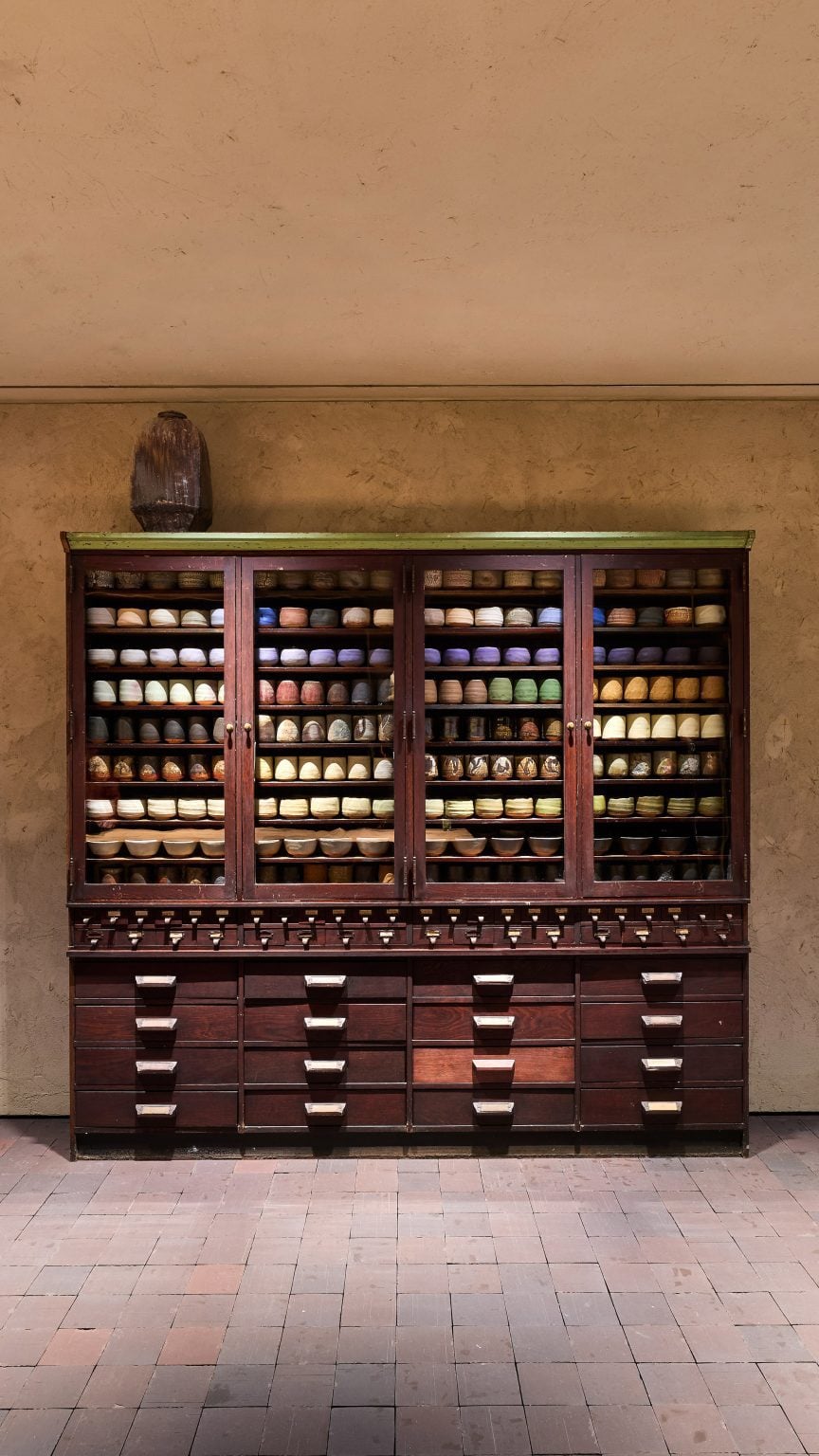 Photo of an antique apothecary cabinet in dark wood holding dozens of similar cups and bowls, with a gentle rainbow of glazes in the Chawan Cabinet exhibition at Prada Home Milan