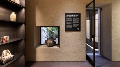 Photo of the Chawan Cabinet exhibition at Prada Home showing a wall hewn with earthy plaster, with a niche cut out in the wall holding a heavy vessel and showing glimpses of a leafy courtyard behind