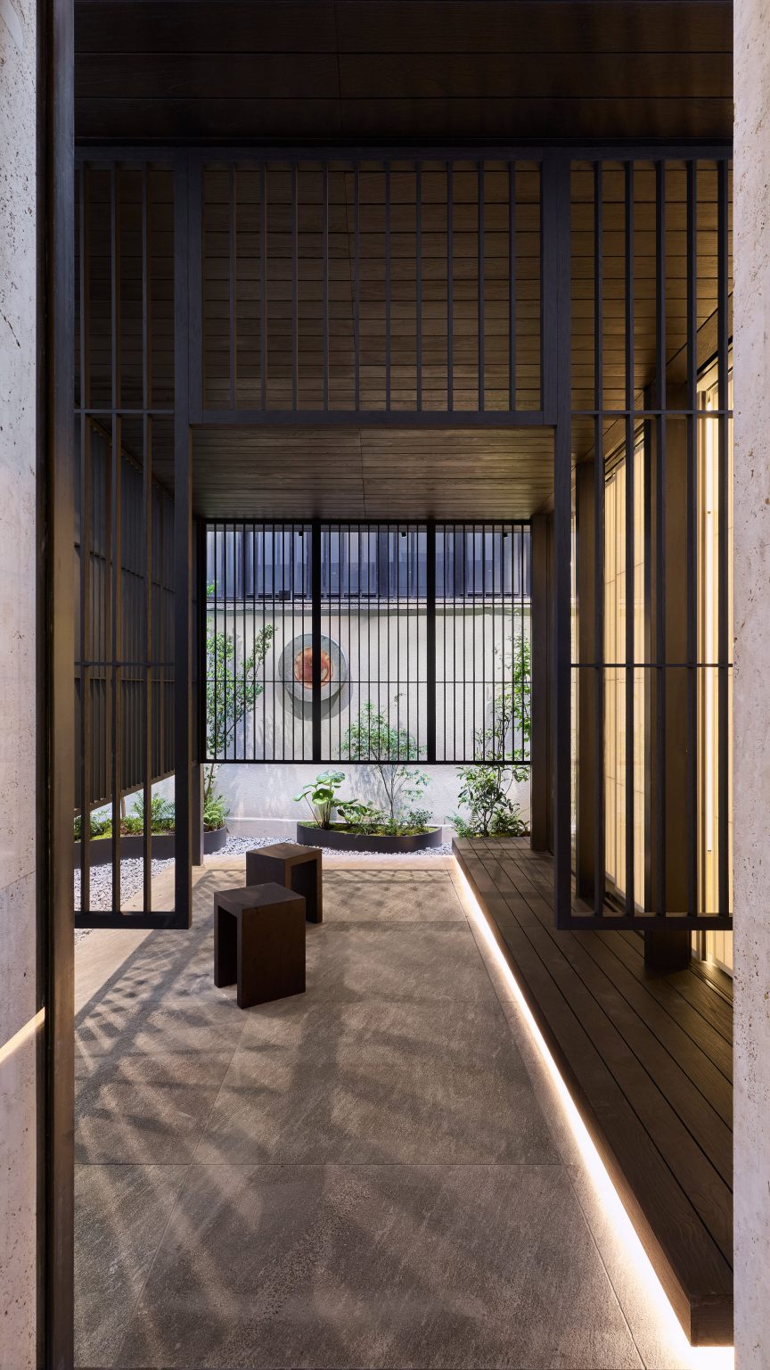 A photo of the courtyard at Prada Home Milan showing a calming space with screens providing seclusion, simple wooden stools, hidden strip lighting and a garden full of pebbles and soft greenery