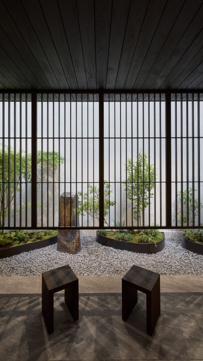 Photo of a calm courtyard decorated with two simple wooden stools and a pebble-filled garden with planters holding small trees and ferns