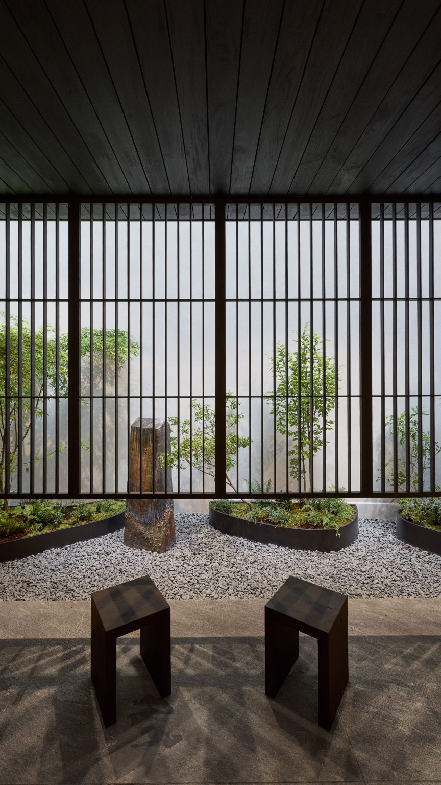 Photo of a calm courtyard decorated with two simple wooden stools and a pebble-filled garden with planters holding small trees and ferns