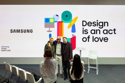 Three men pose on stage in front of a "Design is an act of love" screen as audience members take photos