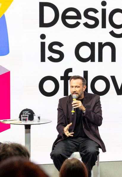 Mauro Porcini speaking into a microphone while seated on stage in front of a "Design is an act of love" backdrop