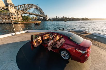 Photo of the Sunburnt Car, a red sedan, with its doors wide open showing its skin-like interior, parked on Sydney's Circular Quay in front of a view of the Sydney Harbour Bridge
