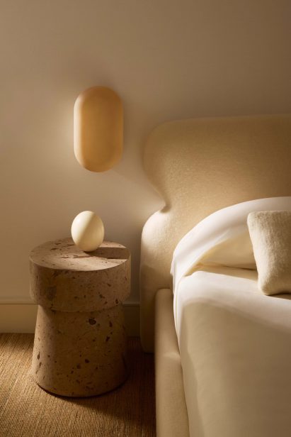 Neutral toned bedroom vignette with a lozenge-shaped sconce, cork side table and sheep wool headboard