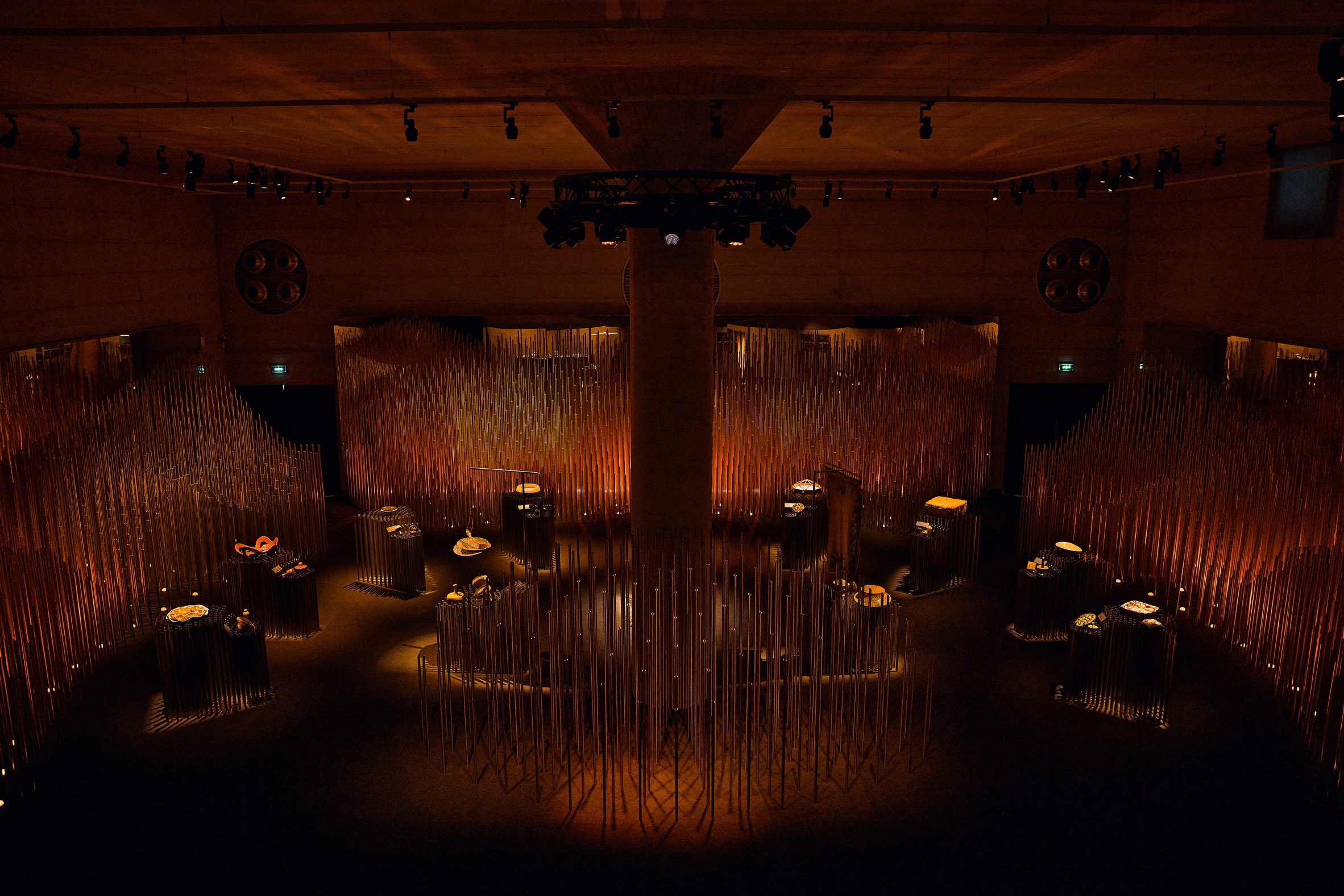 Dimly lit exhibition space with reed-like elements and display plinths showcasing objects