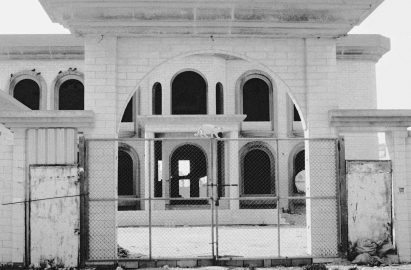 Black and white photograph of a cat on a metal gate, with a large arched building in the background.