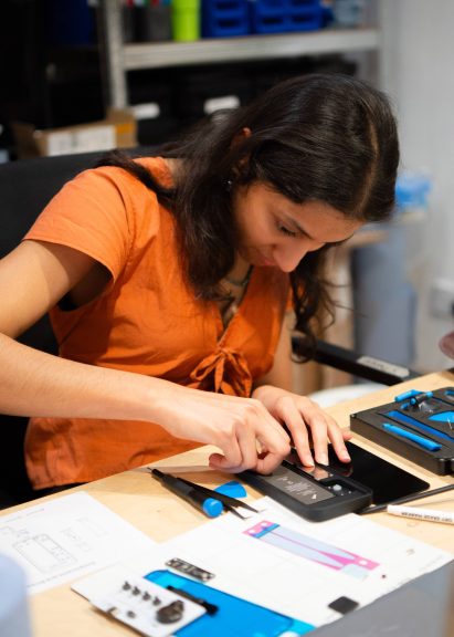 Photo of a woman working at a desk surrounded by bits of electronics as she concentrates on taking apart the inside of a phone