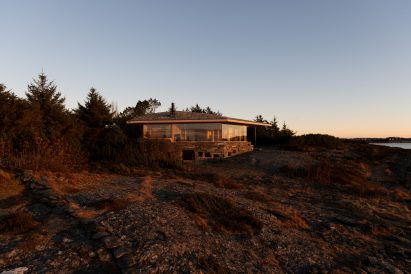 Exterior view of the low timber cabin on a rocky coastal site at sunset