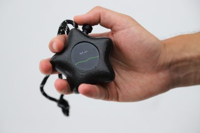 Photo of a hand holding the Mesh-89 device with a slightly wavy green line across a circular black screen with Arabic text above it