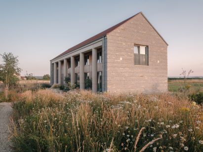 View across meadow grasses towards the rear of the house, showing its pitched roof, stone walls and lower wing extending into the landscape