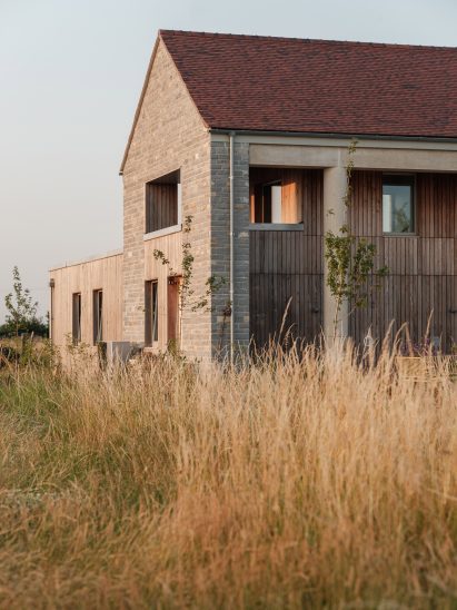 Side view of the farmhouse-style home with pale stone walls, timber cladding and a pitched roof surrounded by long grasses